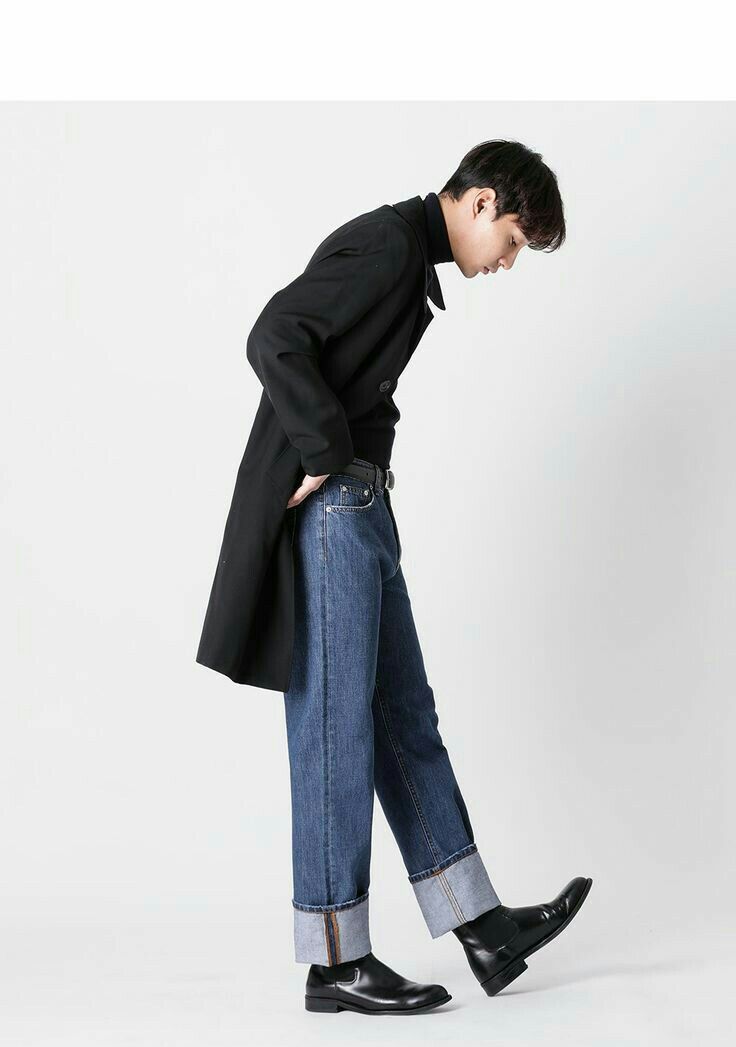 a man in a black coat, blue jeans, and black boots stands in a studio with a white background. he is looking down at the ground. his left foot is forward and his right foot is back. his jeans have been rolled up. he is wearing a black turtleneck shirt.  his hair is black and slicked back.  his coat is long and has a collar. his boots are black leather.  the image is a portrait of a man in fashion clothing.