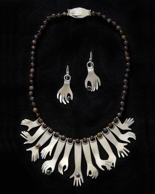 a necklace and earring set are displayed against a black background. the necklace is made of beads with silver hands attached at the bottom. the earrings are silver hands with colored stones in the palms. the necklace also has a silver clasp in the shape of two hands holding each other.