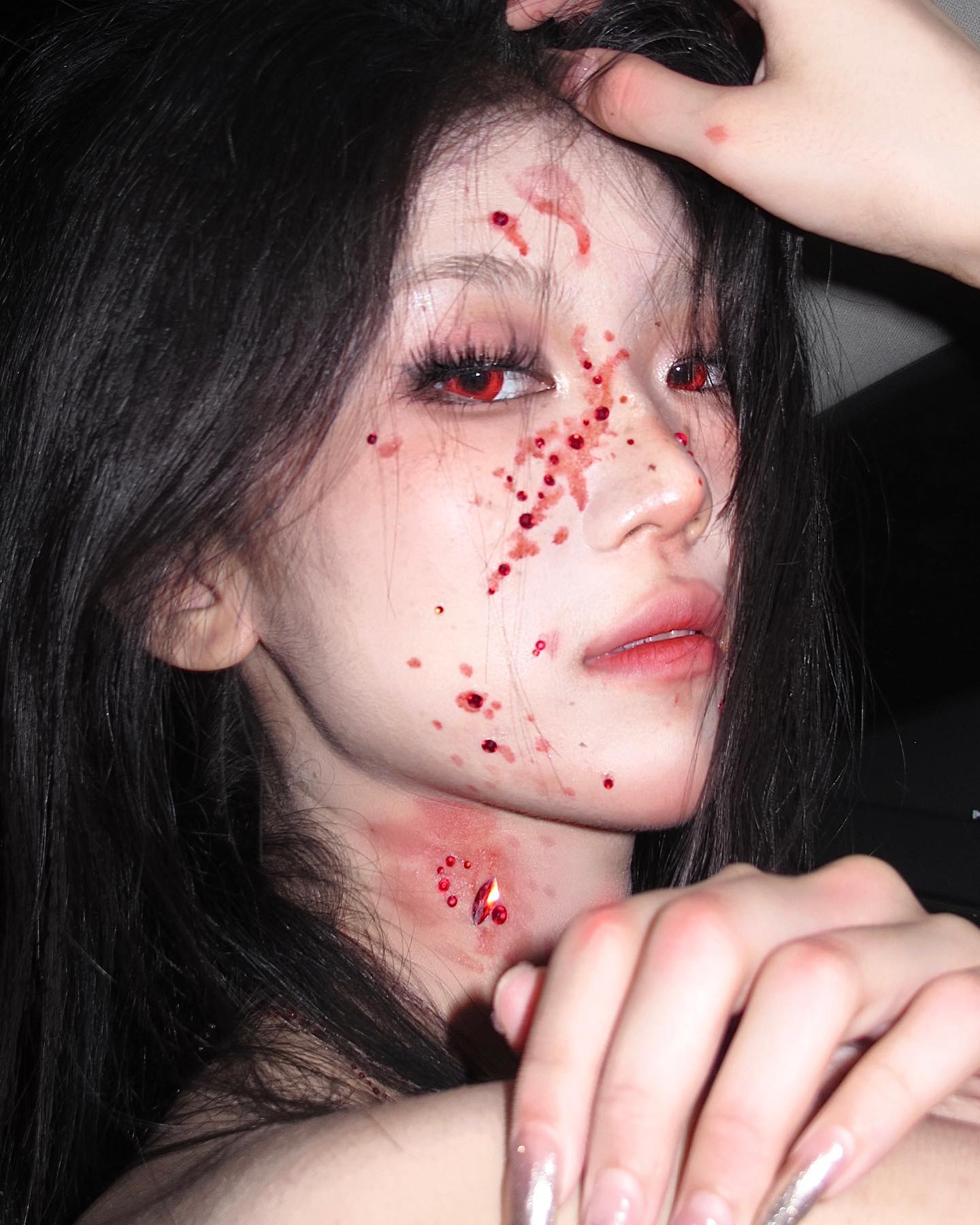 a young woman with long black hair and dramatic makeup poses for a closeup portrait. her look evokes a vampire aesthetic, with fake blood splattered across her face, neck, and chest. she wears red contact lenses and has sparkling red gems adorning her skin. the photo appears to be taken inside a car, with dim, artificial lighting enhancing the dramatic effect. her expression is serious and alluring, highlighting the artistry of her costume and makeup.
