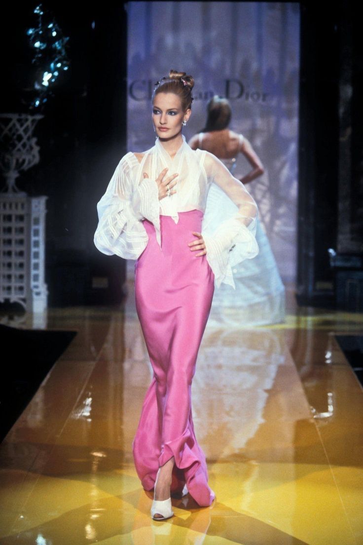 a model walks the runway in a christian dior fashion show. she is wearing a pink, highwaisted dress with a sheer, white blouse. she also has on white shoes and jewelry. the backdrop has the christian dior logo on it and there are lights shining on the stage. another model is visible in the background.