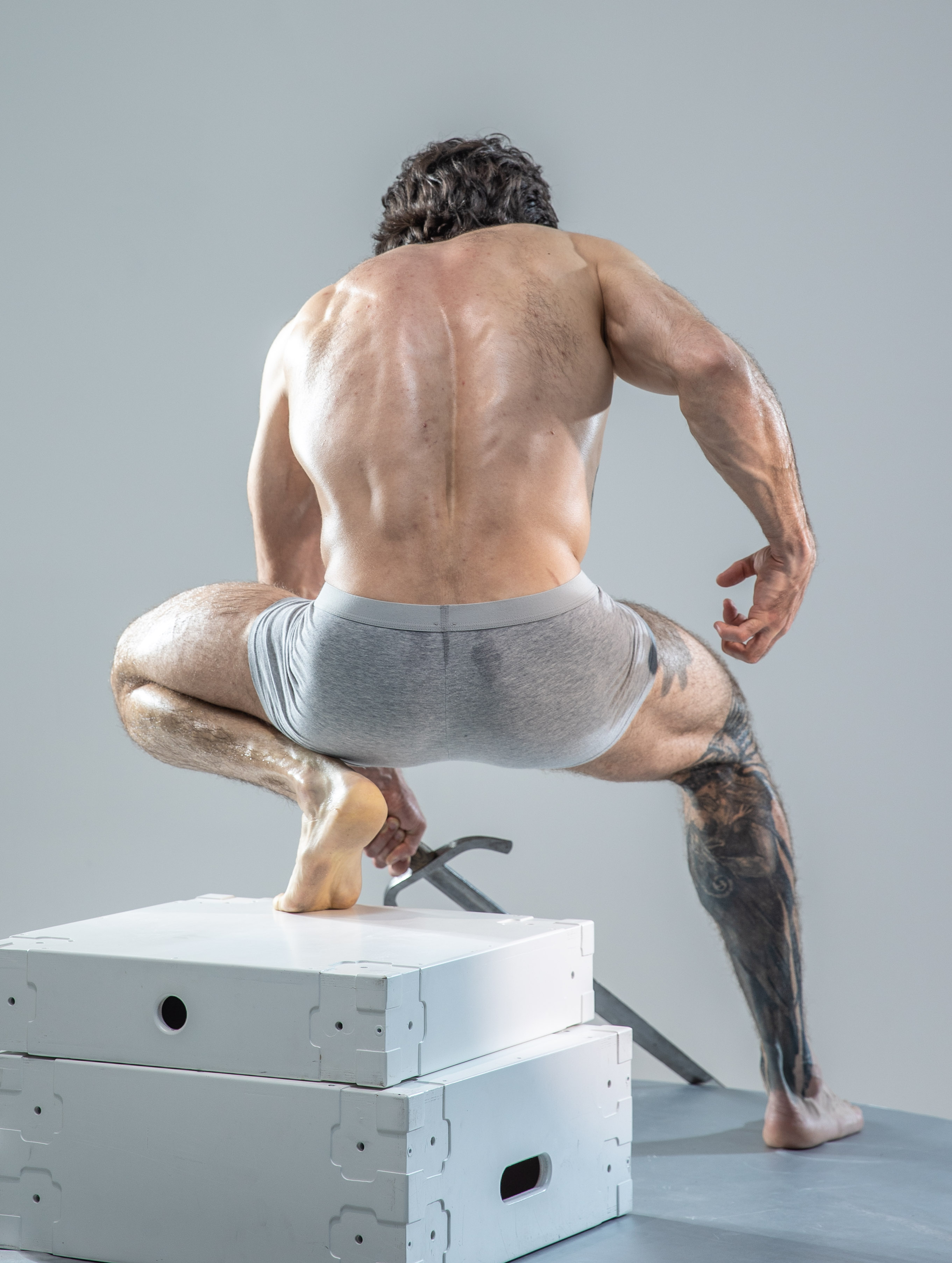 a muscular, athletic man with dark hair is captured from behind, posed in a crouch atop two white storage boxes in a studio setting. he wears gray underwear, showcasing his toned physique. the lighting is soft and diffused, emphasizing the definition of his muscles.