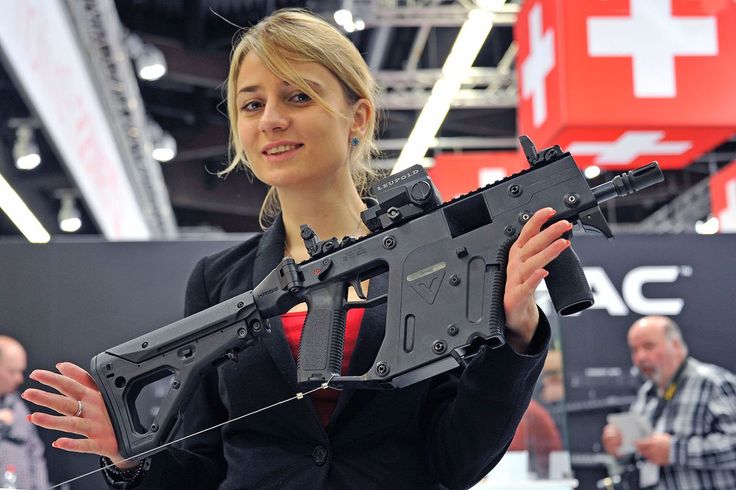 a woman is holding a black kriss vector submachine gun with a leupold red dot sight attached. the background includes a red cube with the swiss flag on it and other people.