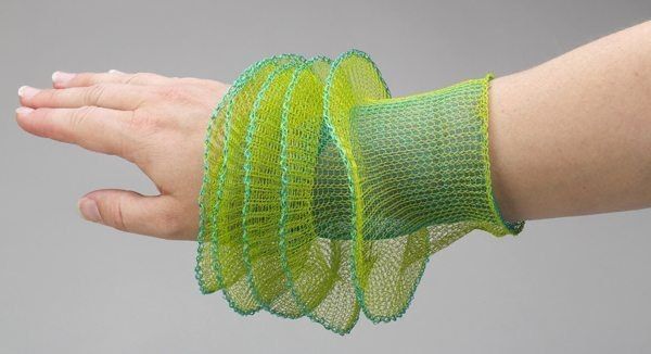 a person's arm and hand are visible, with a bright green and blue mesh bracelet adorning the wrist. the bracelet is large and accordionlike, with ruffled edges. the skin on the hand and arm is fair. the background is a light gray.