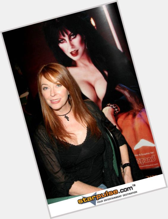 a woman with long red hair and black top is sitting in a chair and smiling at the camera.  a woman with long dark hair and black dress is sitting behind her.  the image is a photo taken from a close up perspective.