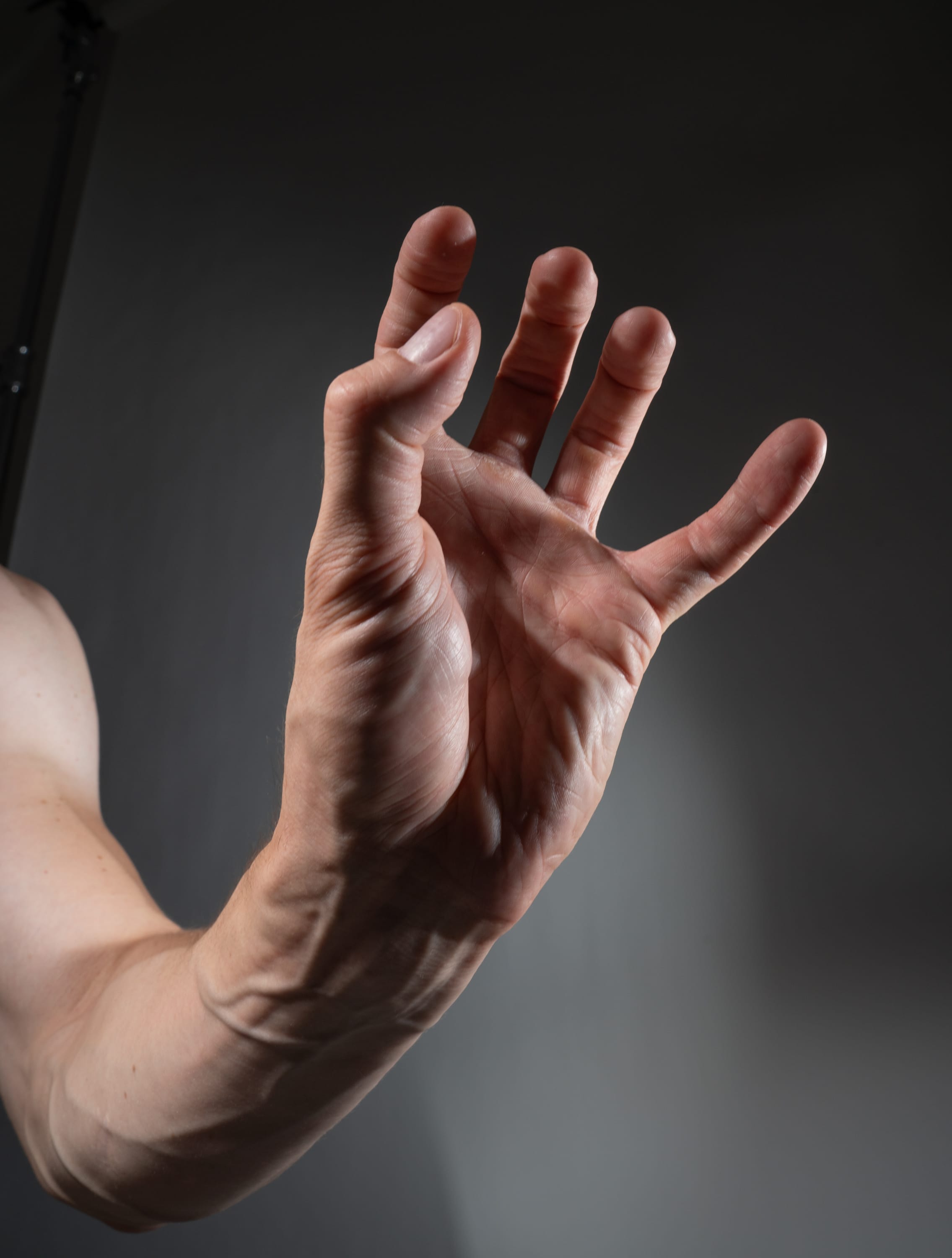a closeup image of a man's outstretched hand and arm against a grey studio background. the hand is positioned with fingers spread, illuminated by studio lighting that creates shadows and highlights the details of the skin and muscle structure.