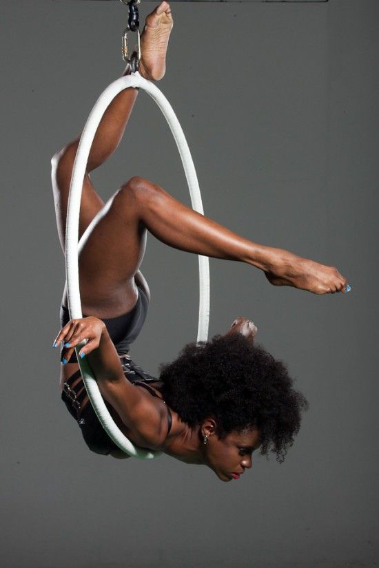 a woman in a black leotard is hanging upside down in an aerial hoop, with her back facing the camera. her legs are bent at the knee and her left leg is wrapped around the hoop. her right foot is hanging straight out, pointing at the camera. she is leaning into the hoop with her left hand, and her right arm is extended out to the side, fingers pointing down. her head is tilted to the side, and her long, curly black hair is falling over her face.  the background is a gray wall.