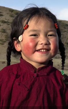 a young asian girl, possibly tibetan, with rosy cheeks and a wide, happy smile, is wearing a traditional red dress and has her hair braided with colorful beads and a shell earring. she is standing outdoors against a backdrop of mountains and sky.