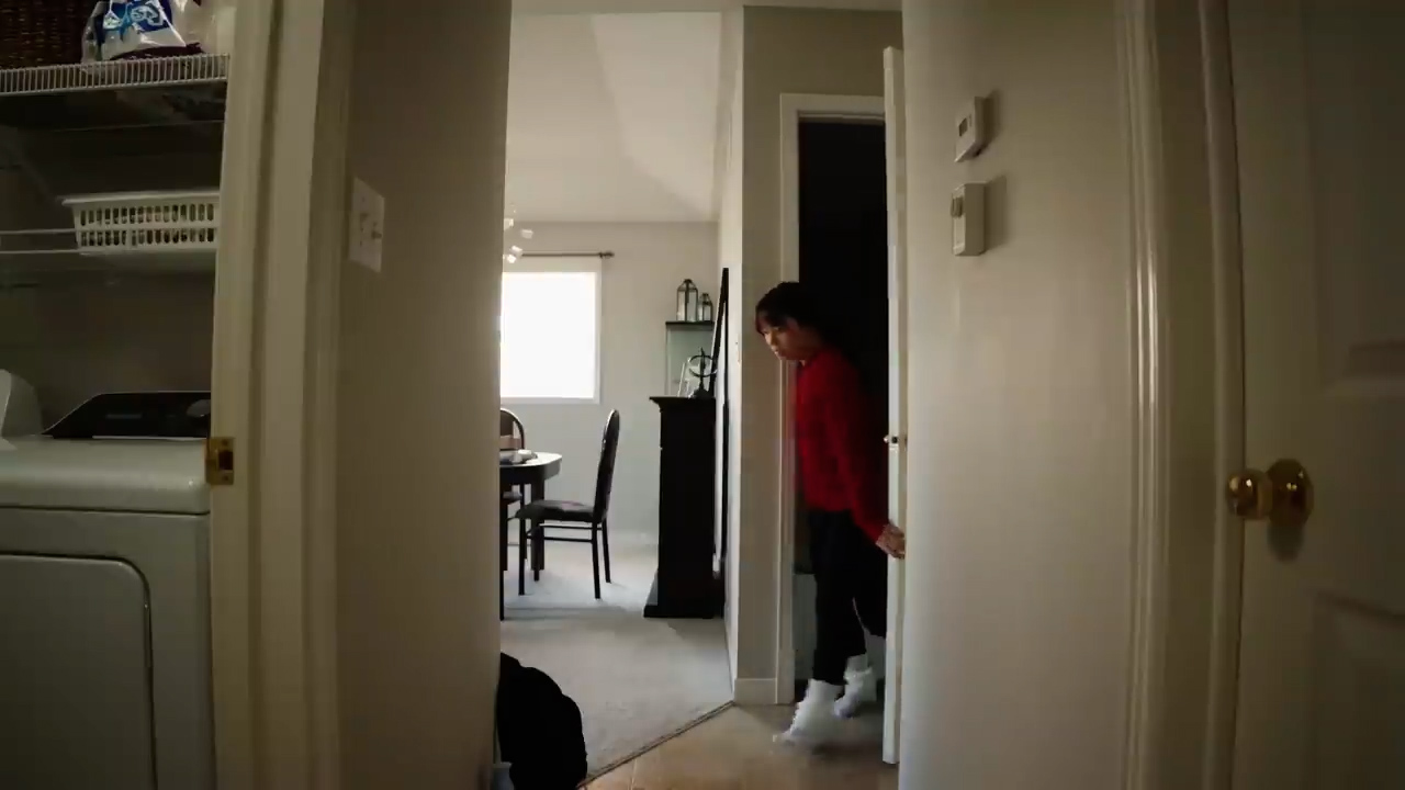 a person wearing red longsleeved shirt and white pants is walking from a laundry room into a hallway. the laundry room has a washer and dryer, and a shelf. the hallway leads to a dining room with a table and chairs. the walls are painted in light gray and the floor is carpeted in light beige.