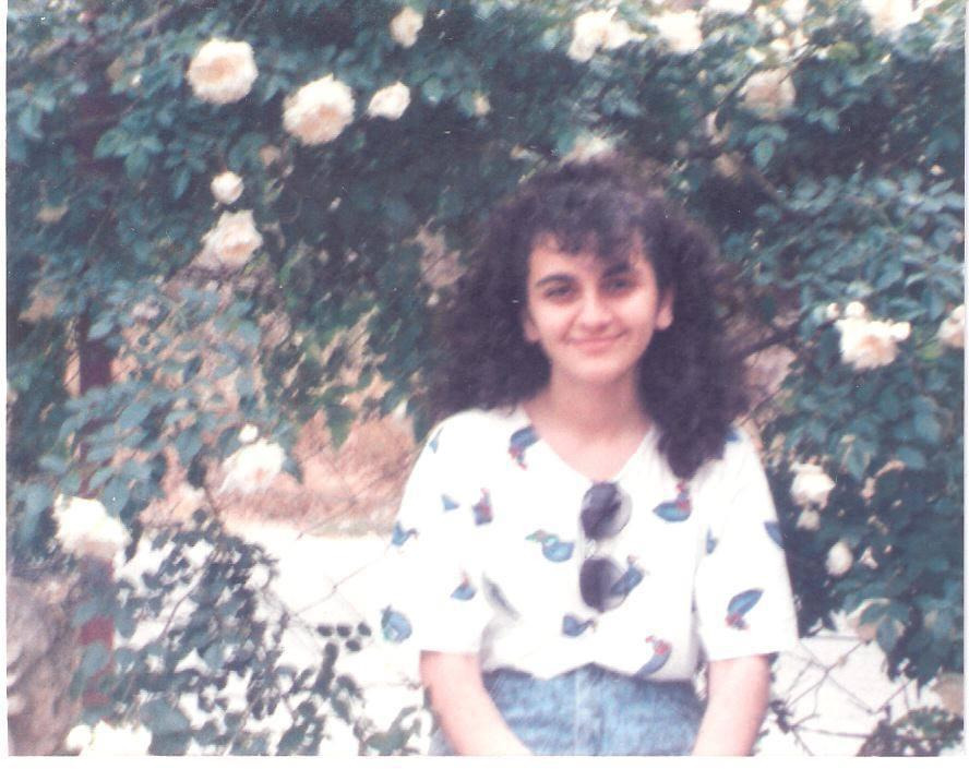 the image is a vintagestyle photograph of a young woman, likely a teenager in the 1990s, sitting outdoors amidst a flowering rose bush. she has dark, curly hair and is smiling pleasantly at the camera. she's wearing a white shortsleeved shirt with a small, repeating bird design and denim shorts. the roses are predominantly white, and the overall feel of the photo is soft and nostalgic, with a slightly faded and blurry quality consistent with older photographs.  the background is lush with greenery and the focus is primarily on the woman.  a pair of sunglasses rests on her shirt.