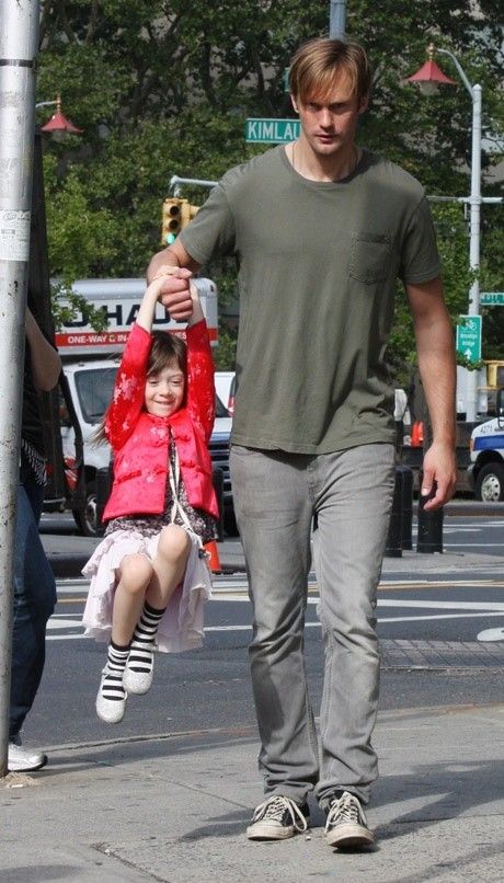 a man with long hair is walking down a city street with his young daughter who is wearing a bright red jacket, holding hands and smiling. the man is wearing jeans and a gray tshirt.  the photo appears to have been taken on a sunny day.