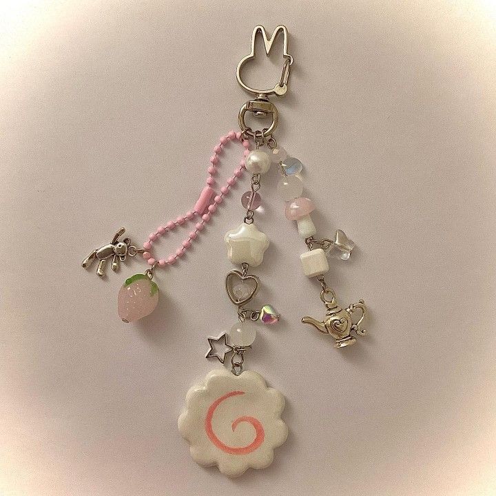 the image shows a kawaiistyle keychain with various charms attached to it. the keychain features a pink beaded chain, along with charms like a strawberry, a rabbit, a star, a heart, a mushroom, and a lamp. the overall color scheme is soft and pastel, with pink and white tones dominating. at the bottom of the keychain is a white cloudshaped charm with a pink spiral, resembling a narutomaki, a type of japanese fish cake. the keychain is placed against a neutral background.