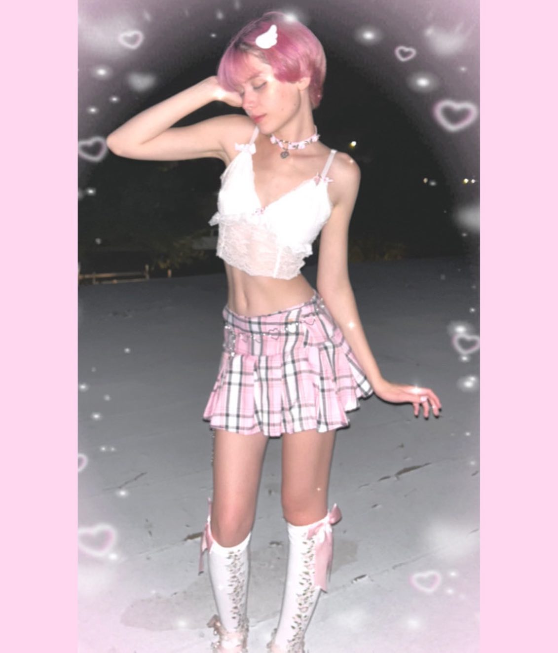 the image shows a young woman with short, bright pink hair. she's wearing a white lace bralette, a pink and white plaid pleated mini skirt, and white kneehigh socks with pink bows.  she has on a delicate choker necklace. the setting appears to be outdoors at night. the image is heavily edited with a filter that adds hearts and a soft, dreamy effect. the overall style is consistent with pastel goth or kawaii fashion aesthetics.