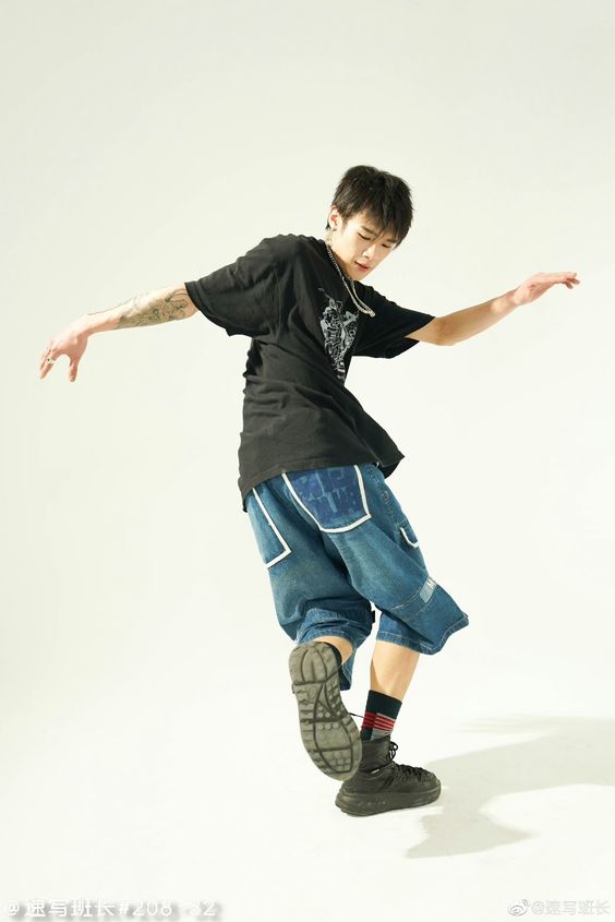 a young chinese man dressed in a black tshirt, blue denim pants, and black sneakers is posing for a studio photo shoot. he is  standing in a relaxed pose, his left arm is raised up while his right arm is bent, and he is looking down at the floor. the image has a white background.
