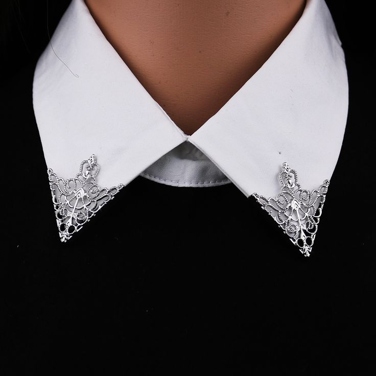 loseup view of a silver, ornate collar pin affixed to the corners of a white shirt collar. the pin is triangular in shape with a lacy, filigree design.  the collar is worn by a mannequin, and the background is black. the overall style is elegant and vintageinspired.