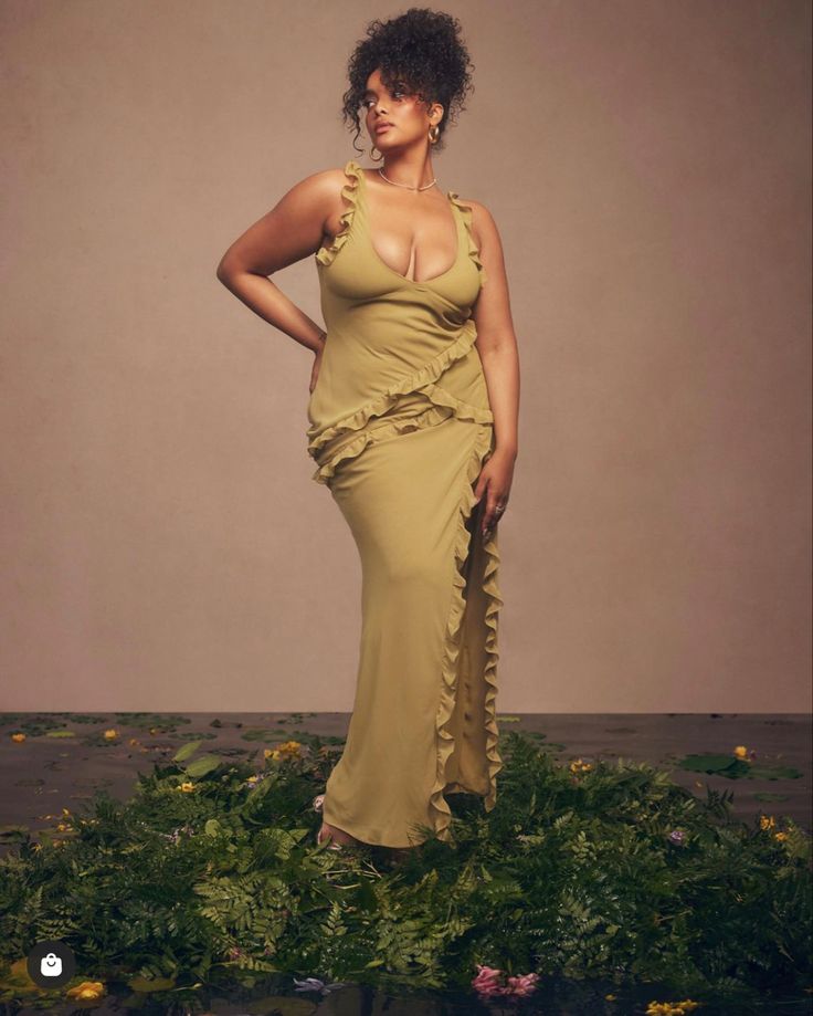 the image showcases a plussize model with dark curly hair. she is wearing a flowing, olive green maxi dress that features ruffles along the neckline and a side slit. the dress is styled in a wrap fashion. the model is standing amidst a bed of lush green foliage and scattered flowers, creating a natural, somewhat mystical ambiance. the background is a muted, neutral tone, making the model and her dress the focal point. the overall aesthetic is one of elegance, natural beauty, and body positivity. the photo seems to be a professional studio shot, promoting either plussize fashion or the specific dress.