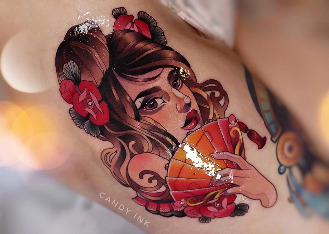a closeup photograph showcasing a vibrant tattoo of a woman on someone's skin. the woman in the tattoo has brown hair adorned with red flowers and holds a red and gold fan. the tattoo is executed in a traditional style with bold lines and a rich color palette.