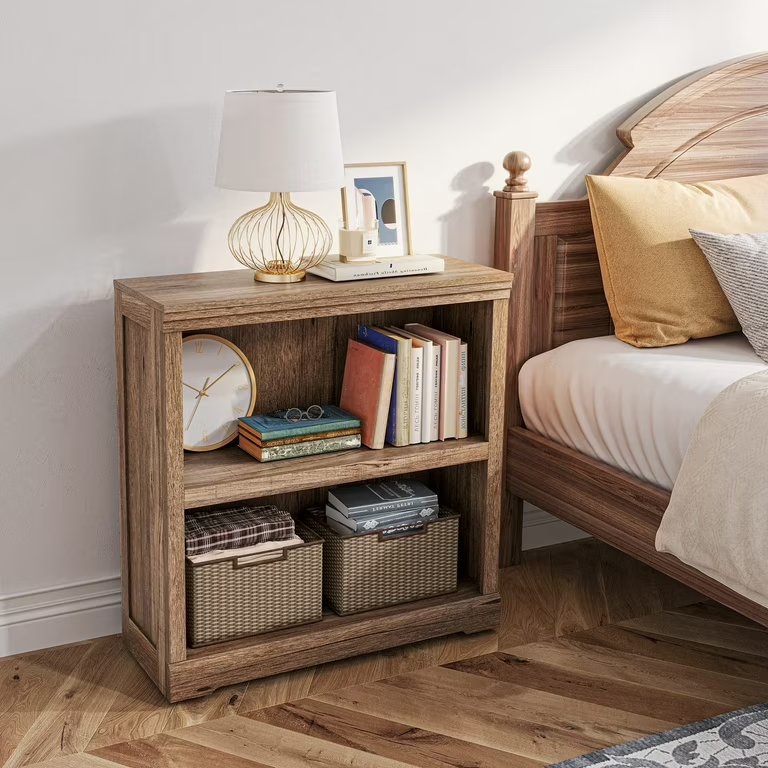 the image showcases a rustic brown wooden bookcase in a bedroom setting. it features two open shelves stacked vertically, providing ample storage space. the bookcase is styled with a collection of books, two woven baskets, a clock, a pair of glasses, and decorative items. a lamp and a picture frame are placed on top of the bookcase, adding to the cozy and personalized ambiance. the bookcase is positioned next to a bed with a wooden headboard, complementing the overall bedroom decor. the room is welllit with natural light streaming in.