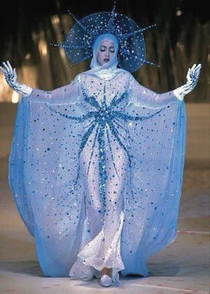 madonna is standing on a runway in a blue sheer gown with a blue snowflake design, a cloak covered in glitter, white gloves, a headdress, and a blue halo behind her head.