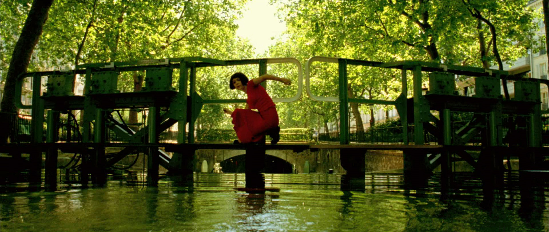 a woman in a red dress dances on a green bridge over a canal in paris. the trees are green and the water is calm. the scene is bright and sunny and the woman looks happy and carefree.