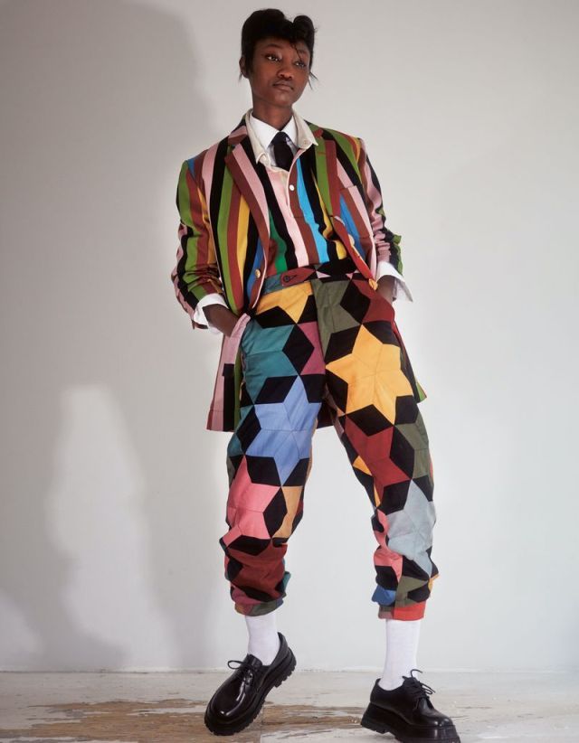 a young fashion model is standing with their feet spread apart. they are wearing a very colorful patterned pants suit and have a dark tie as an accent. their shoes are also black.