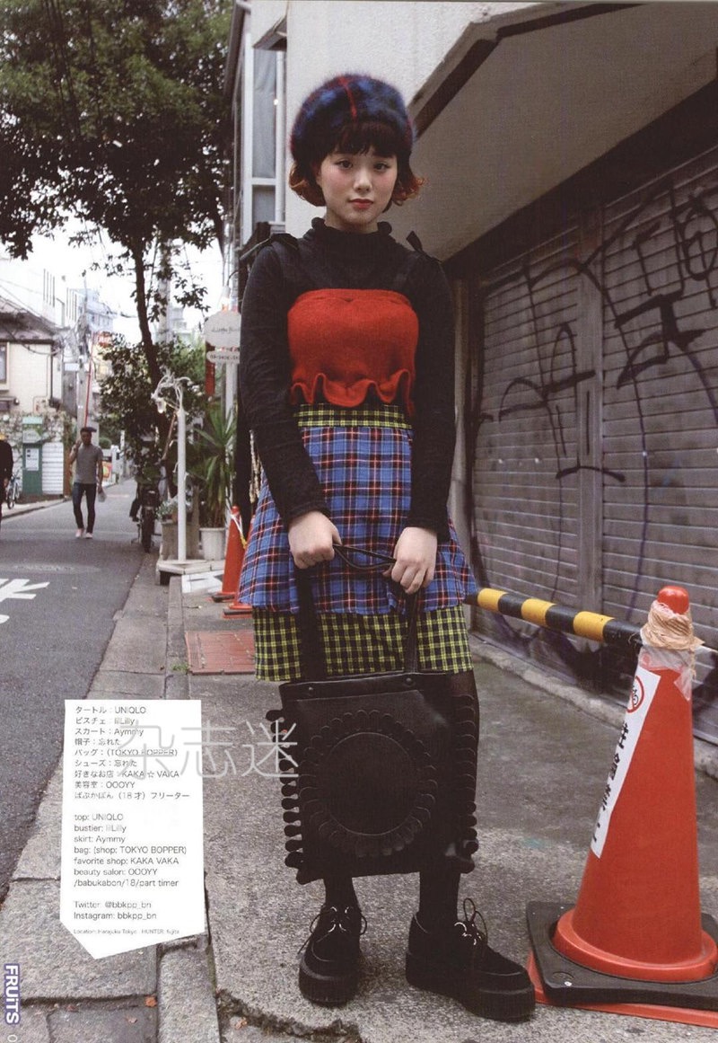 a young woman poses on a city street, showcasing a stylish and eclectic outfit. she wears a red cropped top, a plaid skirt with a layered hemline, and a black beanie hat. her accessories include a black tote bag with a unique design and black platform shoes. the photo captures a sense of urban fashion and individuality.