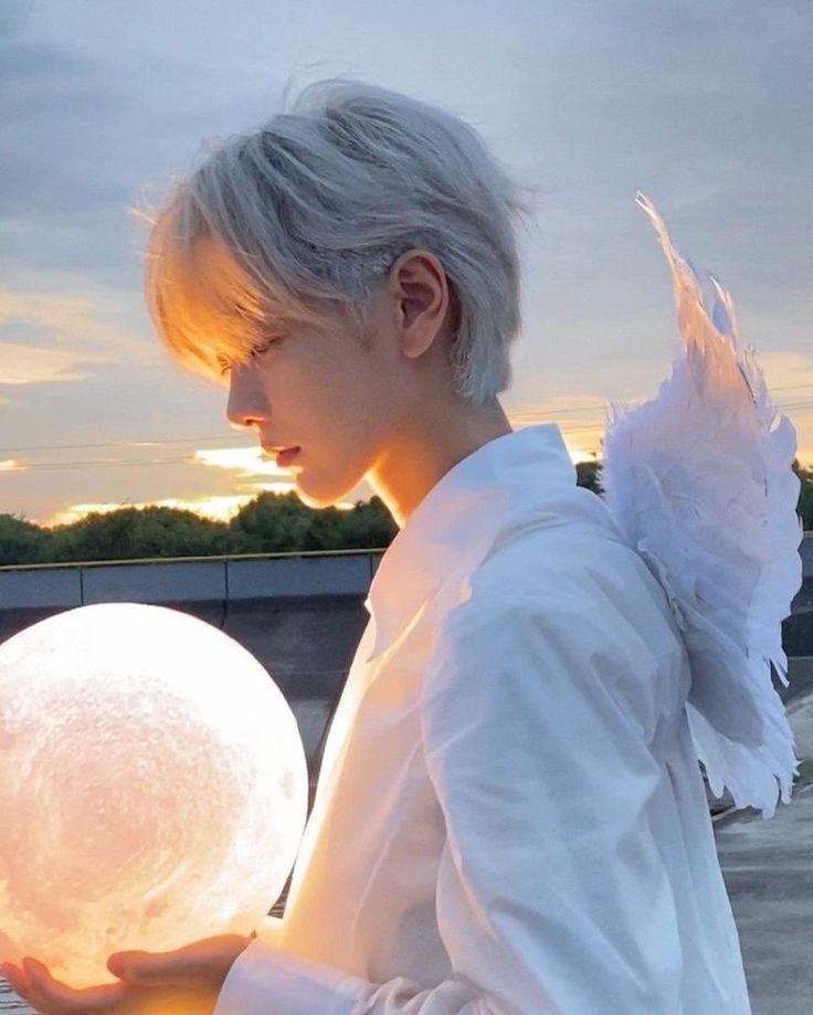 a young man with silver hair and white shirt, adorned with white feathery angel wings, stands in profile against a sunset sky. he gently holds a glowing moonshaped object, casting a soft light on his face. the overall ambiance is serene and dreamlike.