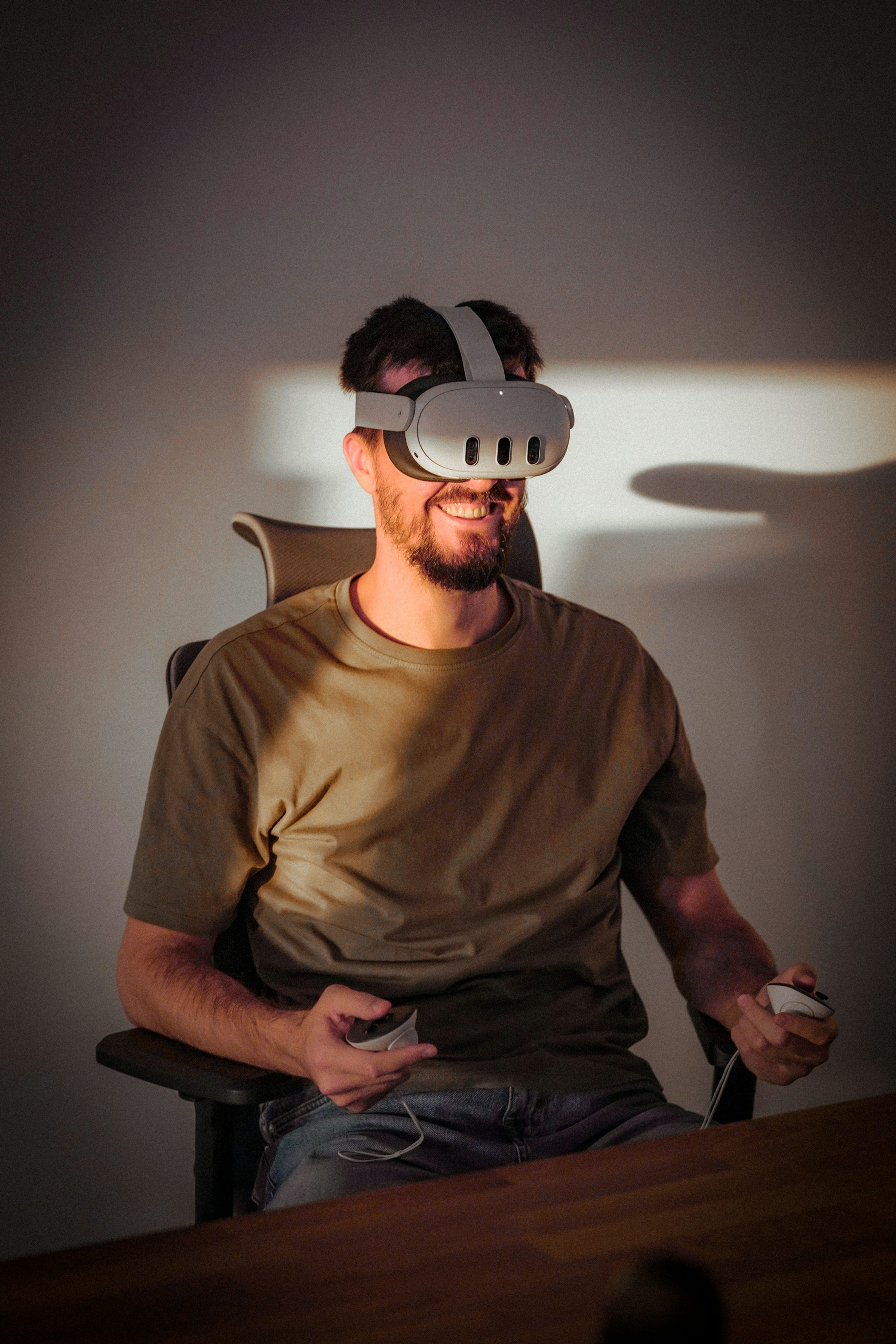 a man is sitting in a chair indoors, wearing a virtual reality (vr) headset and holding controllers. he has a beard and is smiling, suggesting he is enjoying the vr experience. he is wearing casual wear, and the scene is lit with a warm light, creating shadows on the wall.