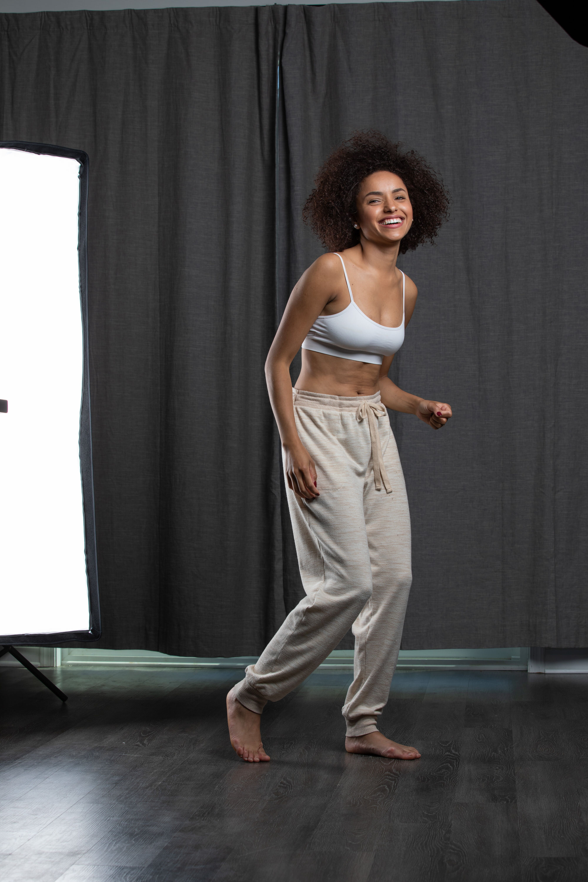 a woman with curly hair is standing in front of a grey curtain backdrop with a softbox on her left. she is wearing a white sports bra and beige joggers. she is smiling and looking happy.