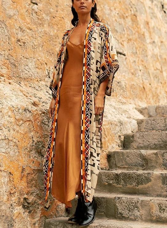 a woman in a long brown dress and patterned kimono stands on stone steps next to a stone wall. she is wearing black ankle boots.