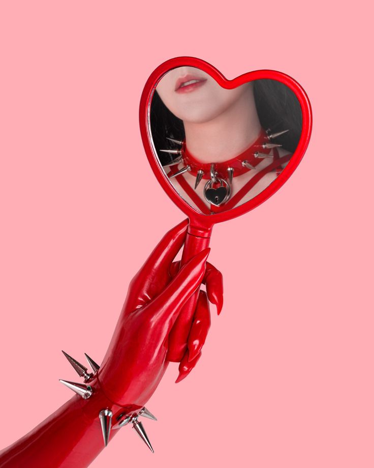 the image is a surreal, stylized composition. a hand wearing a red latex glove with metal spikes holds a heartshaped hand mirror.  the mirror reflects a person wearing a red spiked collar with a padlock. the background is a solid, pale pink.  the overall aesthetic is goth, punk, or alternative, with a hint of fetish or bdsm influence. the contrast between the harshness of the spikes and the softness of the heart shape creates a visually intriguing juxtaposition.