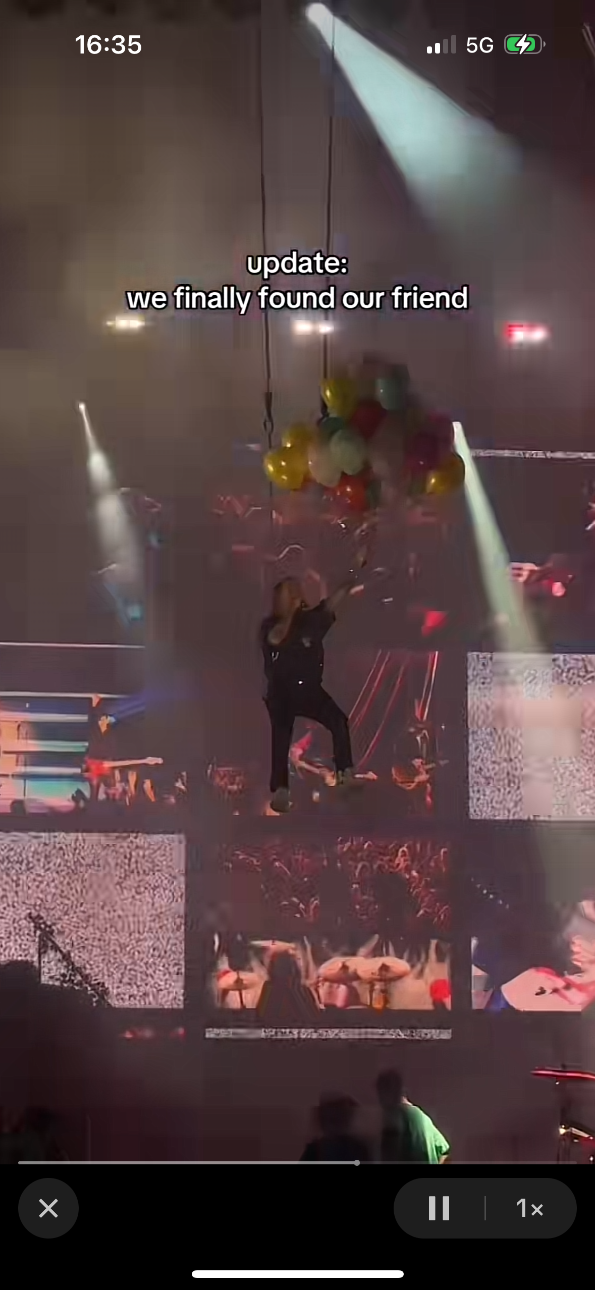 a person is standing on stage with a crowd of people behind them and a banner that reads "update: we finally found our friend".  the stage is lit with bright lights and there are balloons hanging from the ceiling. the people in the crowd are silhouetted against the bright lights. the person on stage appears to be a performer at a concert.