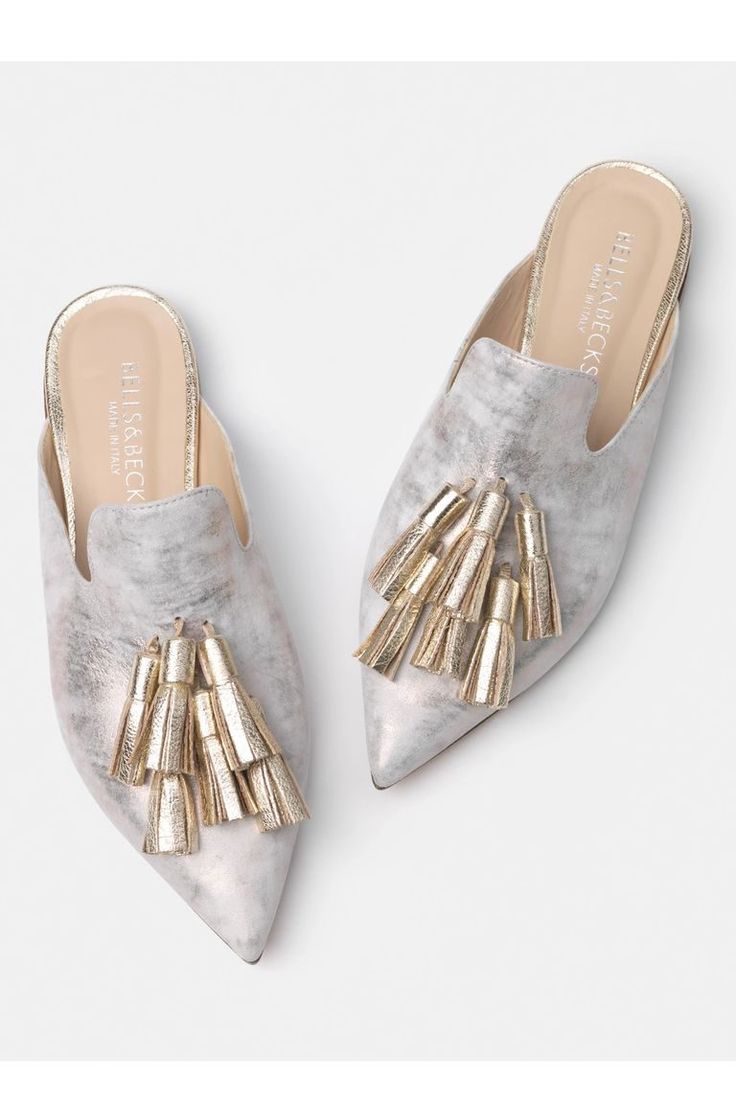 two light grey mules with pointed toes and gold tassel embellishments are displayed against a white background.