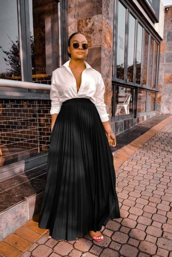 a woman in a white button down shirt and long black pleated skirt walks down a sidewalk outside of a store. she is wearing sunglasses and heels. the sidewalk is made of brick and the storefront windows behind her reflect a city scene.