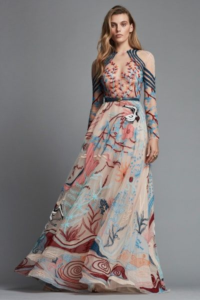 a woman in a long, flowing dress with floral embroidery in pink, blue, and red poses on a gray background. the dress is longsleeved and has a flowing skirt. she is wearing a belt with a buckle at her waist. she has long, wavy hair and a neutral makeup look.