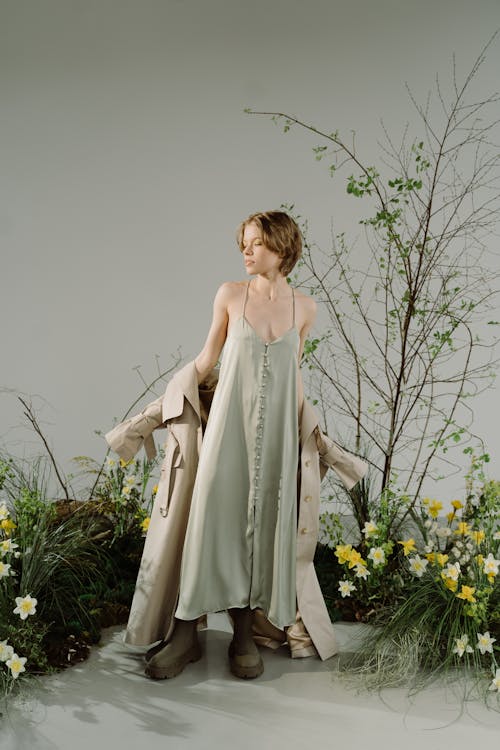 a fullbody shot of a young woman, a fashion model, standing gracefully against a light gray backdrop. she wears a pale sage green maxi dress with delicate buttons running down the front.  she holds a beige trench coat, partially draped around her, and is wearing ankle boots. the background features a naturalistic setting of spring flowers, mainly daffodils, and a leafy, barebranched tree or bush, creating a soft and elegant contrast. the overall style is minimalist and suggestive of a spring fashion shoot. the lighting is soft and natural, enhancing the subtle colours of the clothing and setting.