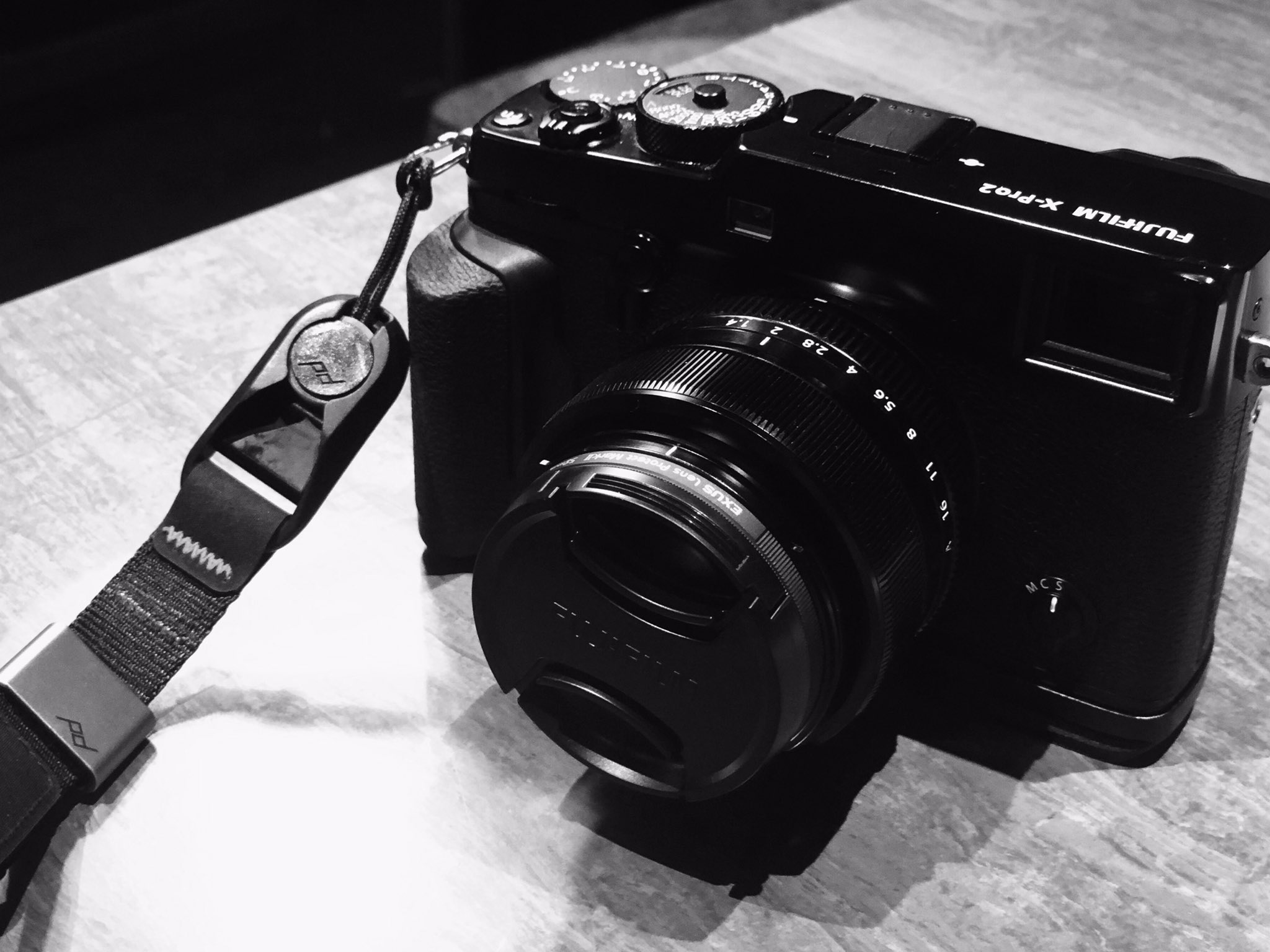 a black and white photo of a fujifilm xpro2 camera resting on a table. the camera is facing the viewer and has a lens cap on. there is a camera strap attached to the camera.