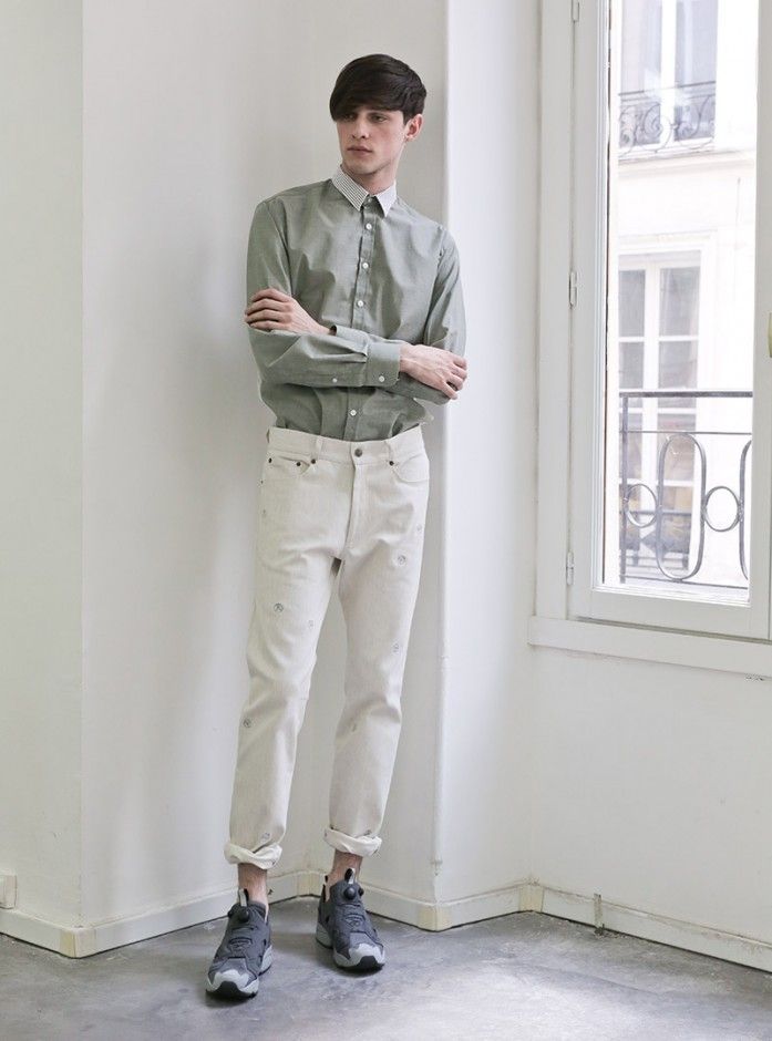 a full shot of a male model standing in a white corner, wearing a green buttondown shirt with a white collar, white jeans with cuffs, and grey sneakers. he has his arms crossed. a window is to the right of him, allowing light into the corner.