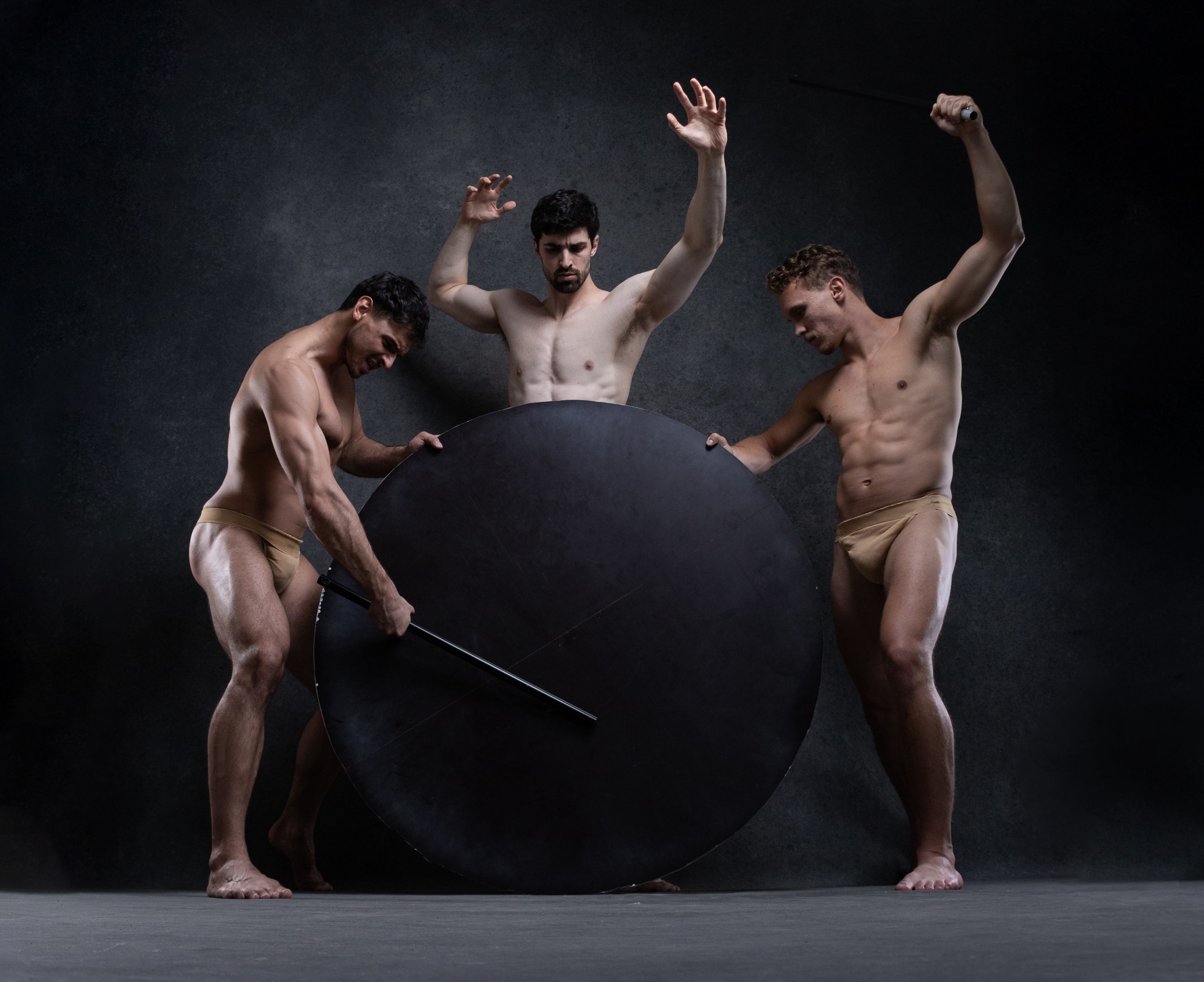 the image features three muscular men in a dramatic pose against a dark, textured background.  they are nude except for small beige briefs. the men surround a large black circle, interacting with it in different ways. one man on the left uses a staff to seemingly push the circle, while another on the right raises a staff above his head as if to strike. the central figure appears caught between the two, his hands raised in a gesture of defense or supplication. the studio lighting emphasizes the contours of their bodies and creates a sense of dynamic tension. the overall effect is one of powerful, yet vulnerable masculinity.