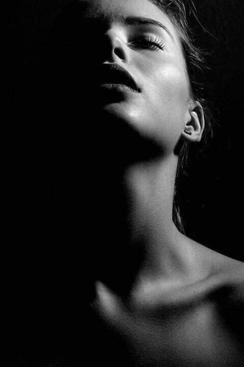 a black and white portrait of a woman. she is lit from the right side of the frame, casting her face and neck into shadow. her lips are slightly