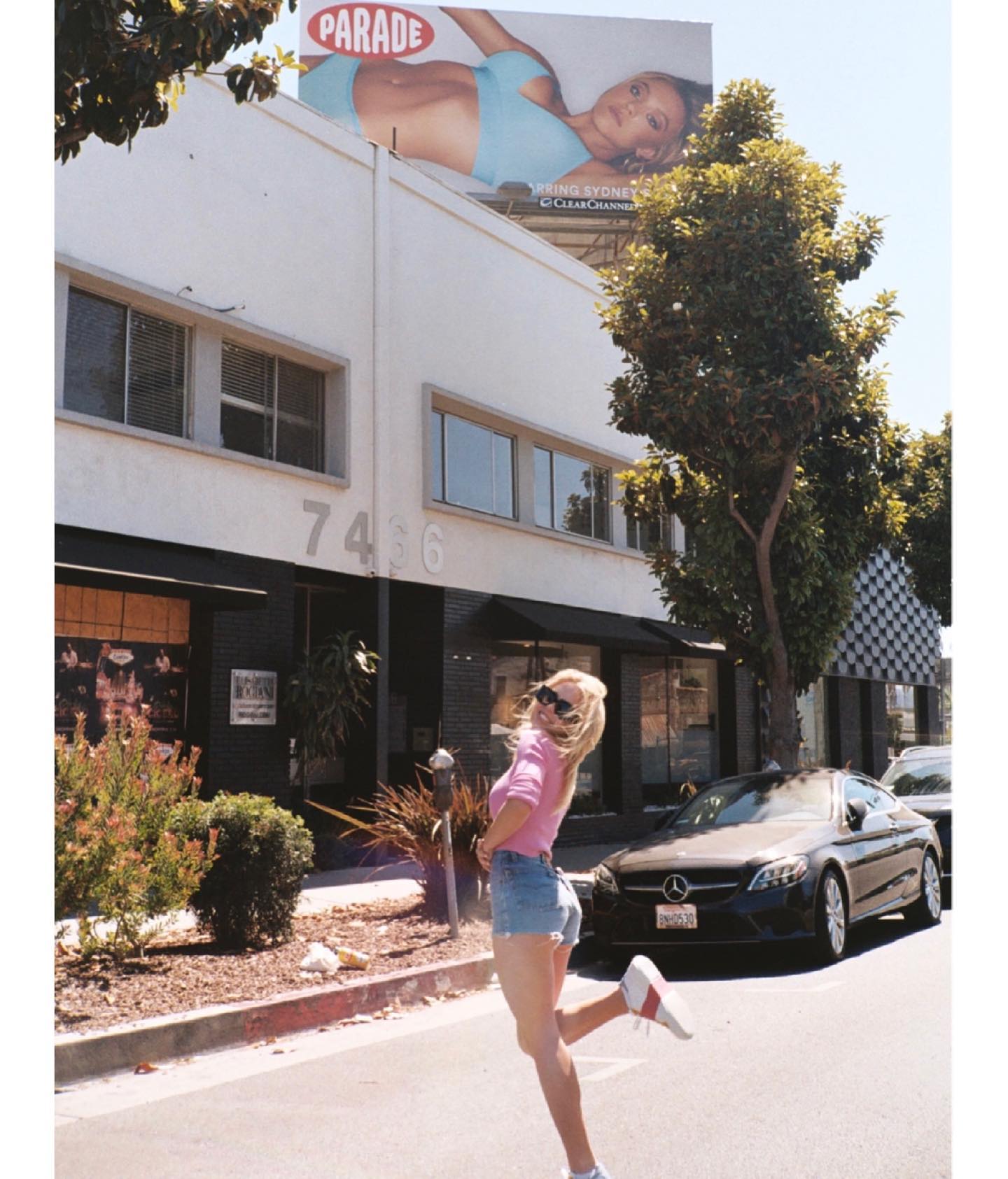 a young woman wearing a pink sweater and denim shorts playfully poses on a city street. she is smiling and wearing white sneakers.  a large billboard advertising the "parade" brand with a model in a bikini is visible in the background above a building. the scene is bright and sunny, typical of a summer day in an urban setting.  the overall feel is cheerful and carefree.
