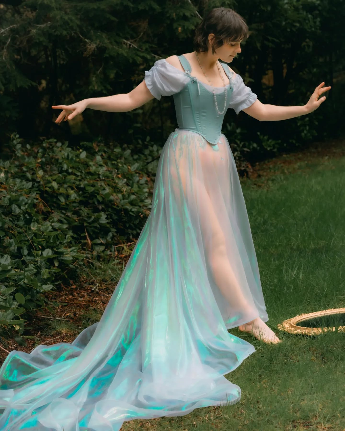 a young woman in a beautiful, flowing pastel green and blue dress poses outdoors in a forest setting. the dress features a sheer overlay and a fitted corset bodice.  the overall aesthetic is ethereal and whimsical. she is barefoot, and a decorative mirror lies on the grass nearby. the photo is styled to look dreamy and romantic.