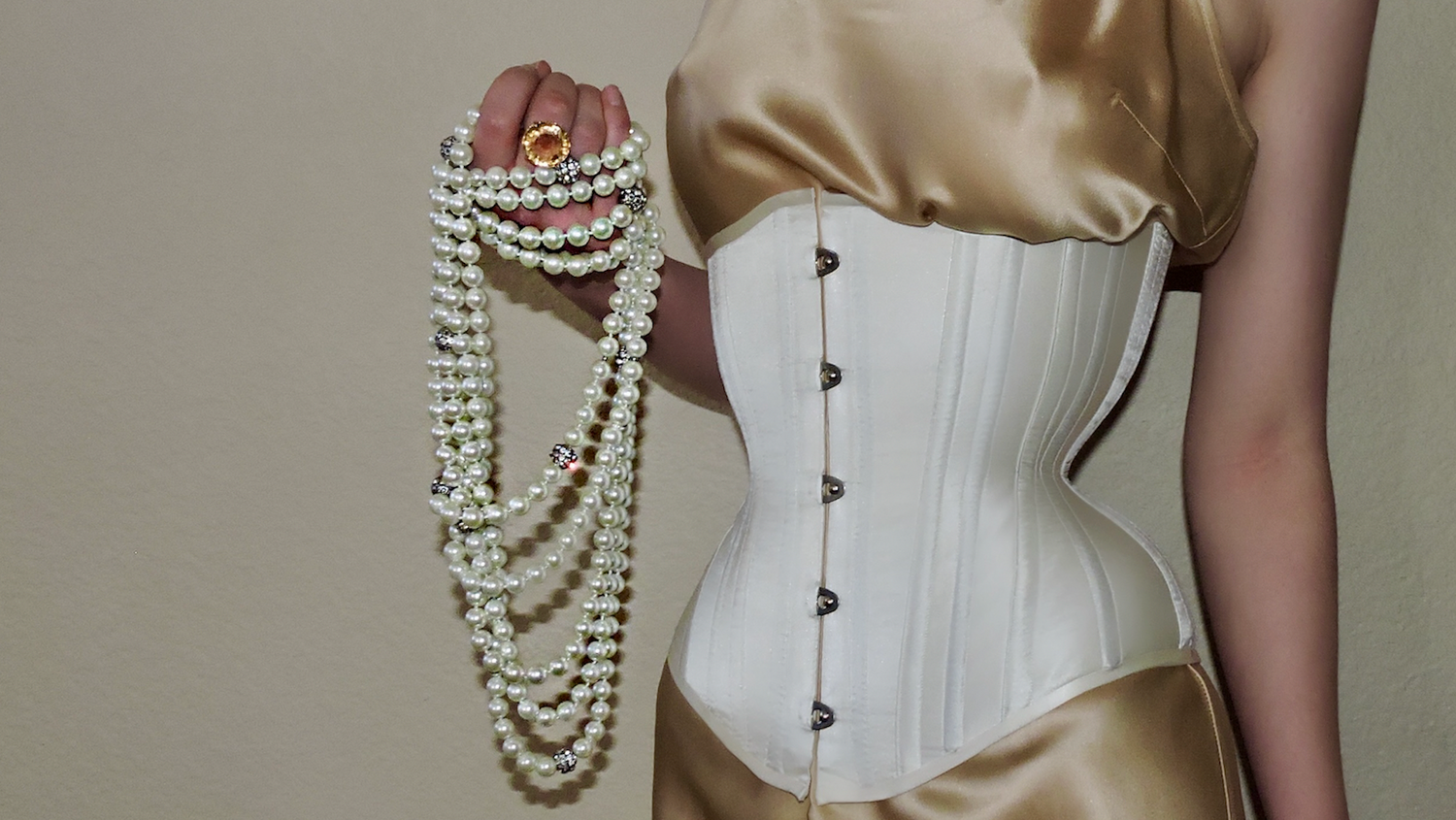 a woman is wearing a white corset and a gold dress, holding a pearl necklace in her hand. the background is a plain wall.