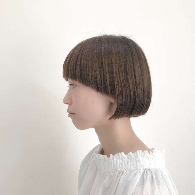 a young asian woman with a short bob haircut. she has straight brown hair with blunt bangs. she is wearing a white shirt and is looking to the side. the background is white.