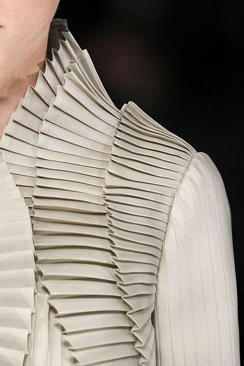 a closeup shot showcases a garment with elaborate pleated fabric detailing. the pleats are arranged in a horizontal fashion, creating a tiered or layered effect around the collar and shoulder area. the garment's fabric has a slightly textured appearance, and the overall design is reminiscent of high fashion or designer clothing, possibly avantgarde in nature. the background is dark, drawing focus to the intricate details of the clothing. the image emphasizes the architectural and textural elements of the garment's design.