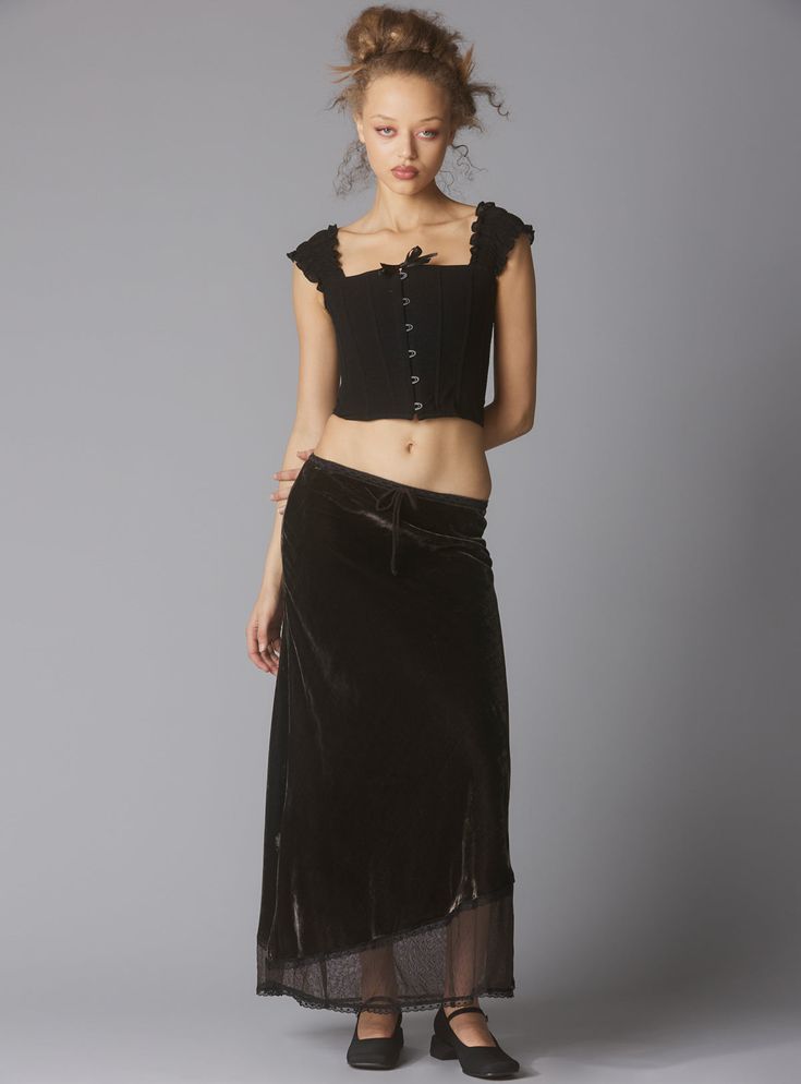 a fullbody shot of a young woman model standing against a neutral grey background. she is wearing a black corsetstyle top with a lace trim and a long, dark brown velvet skirt with a sheer lace trim at the hem. the skirt is styled in a way that drapes slightly asymmetrically.  she has her hair styled in a loose updo with some curls, and she is wearing simple black flats. the overall style is dark, slightly gothic, and vintageinspired.