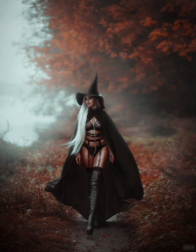 a woman dressed as a witch walks through a forest. she is wearing a black cape, a pointy black hat, and black boots. she is also wearing lingerie and a silver chain around her waist. the forest is covered in autumn leaves. the woman has a mystical, magical and mysterious look.