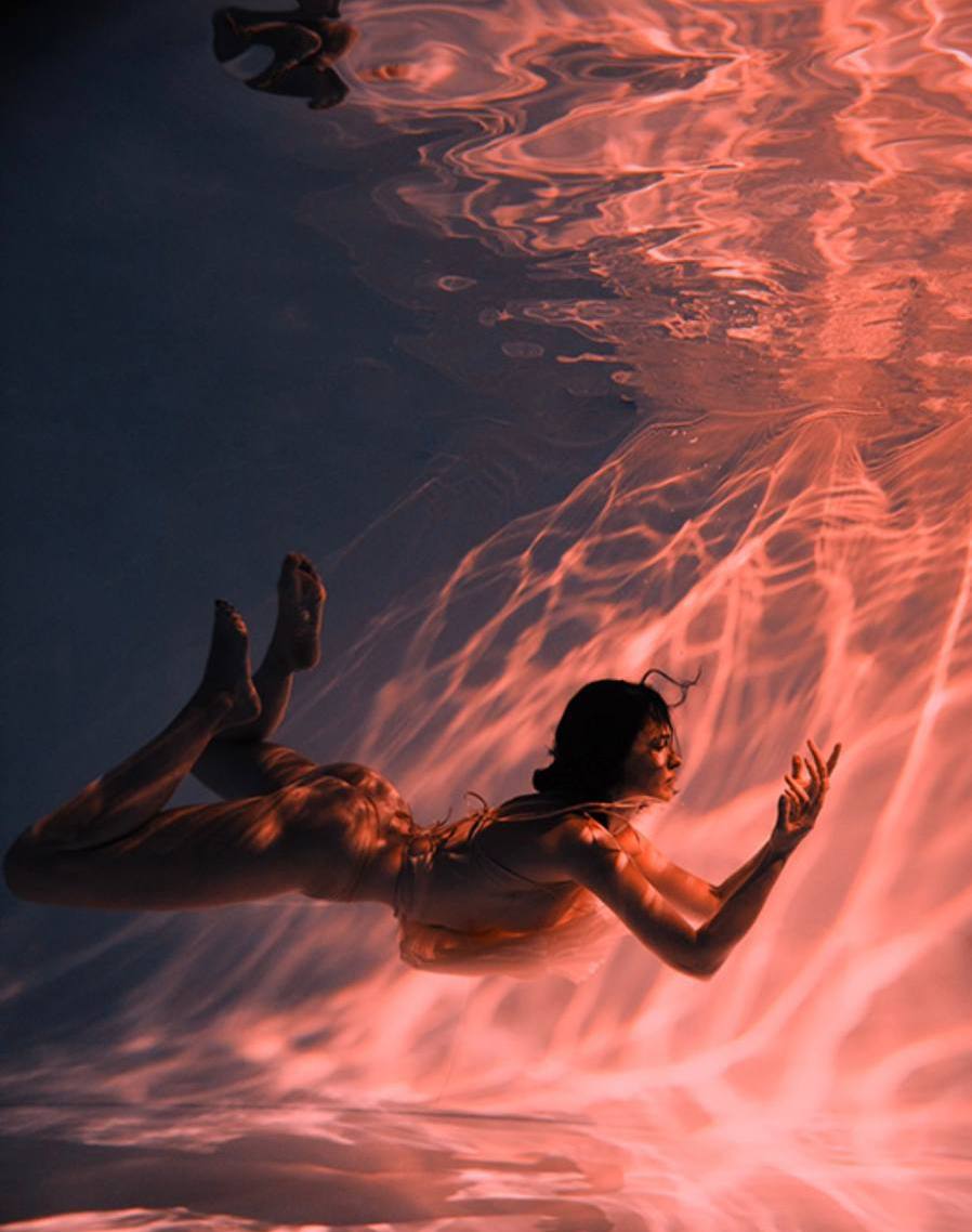 the image is an underwater photograph of a woman gracefully swimming. she is partially submerged, and the water appears to be lit with a warm, orangered hue reminiscent of a sunset. the lighting creates an ethereal and dreamy atmosphere. the woman's body is mostly visible, and she appears to be wearing a sheer, lightcolored garment.  the water itself has a textured appearance with flowing lines and patterns, adding to the artistic feel of the image. the overall mood is serene and captivating.