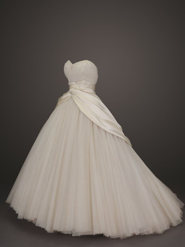 a strapless white wedding dress with a tulle skirt and satin sash draped across the bodice is displayed against a gray background. the dress is a full ballgown shape.