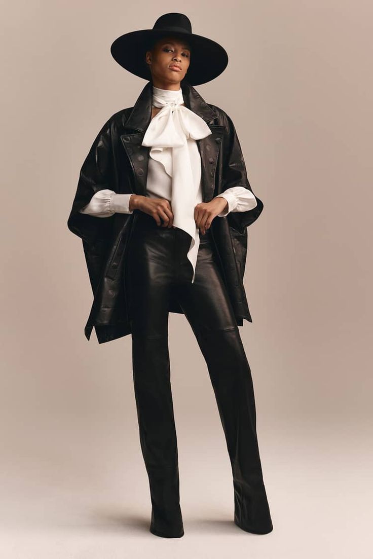 a woman in all black leather outfit with a wide brim hat is posing for the camera. she is wearing a black leather jacket, black leather pants, and a white blouse with a bow. the background is a light beige color.
