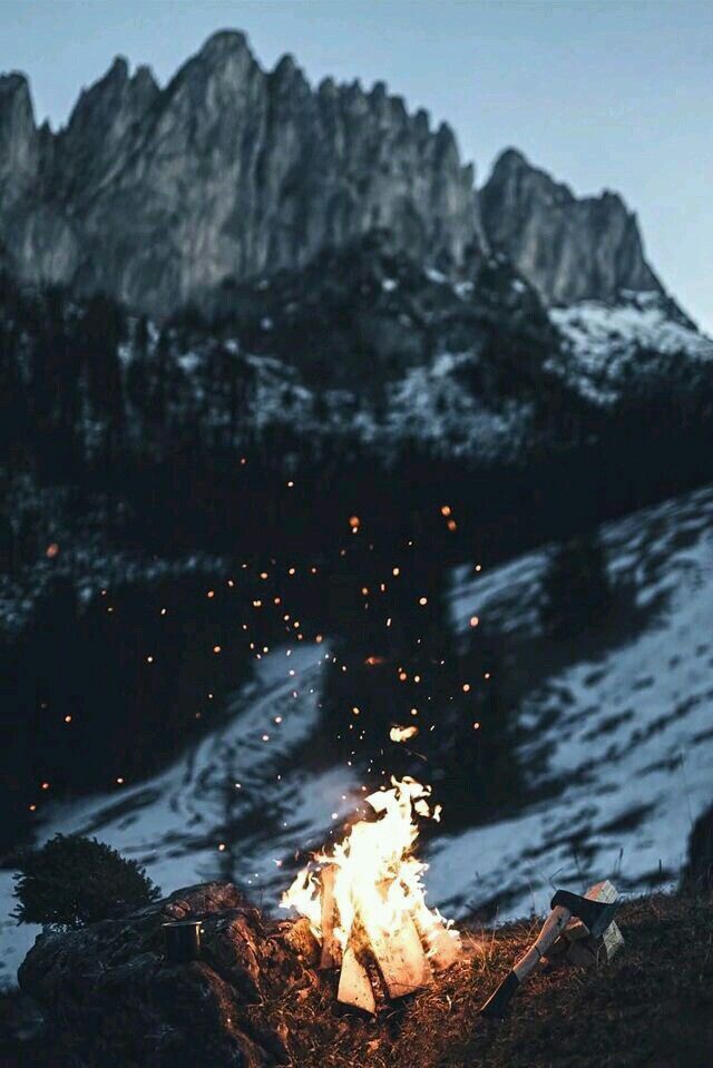 the image shows a campfire burning in a snowy mountain landscape. the fire is surrounded by sparks and an axe is lying on the ground next to it. in the background, there is a dark forest and a tall, jagged mountain range under a dusky sky.